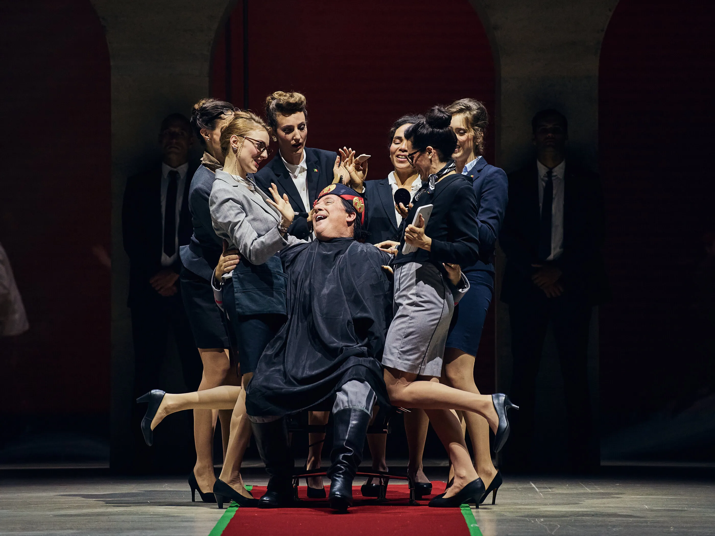 Matthew Brook as Claudio, draped in a barber’s cape and laughing, surrounded by fawning women in business suits