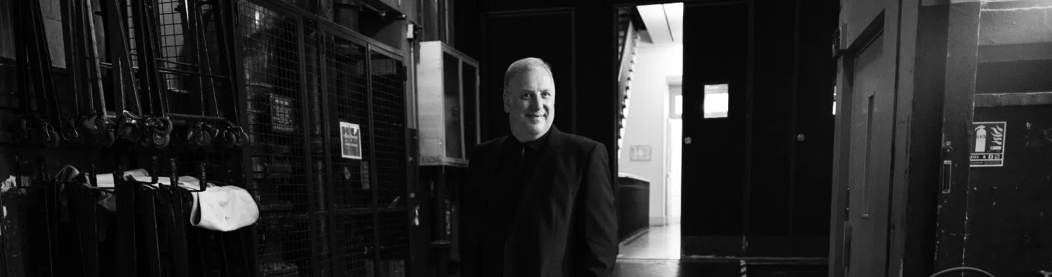 Black-and-white photograph of Matthew Brook backstage, between costume rails and a stage door