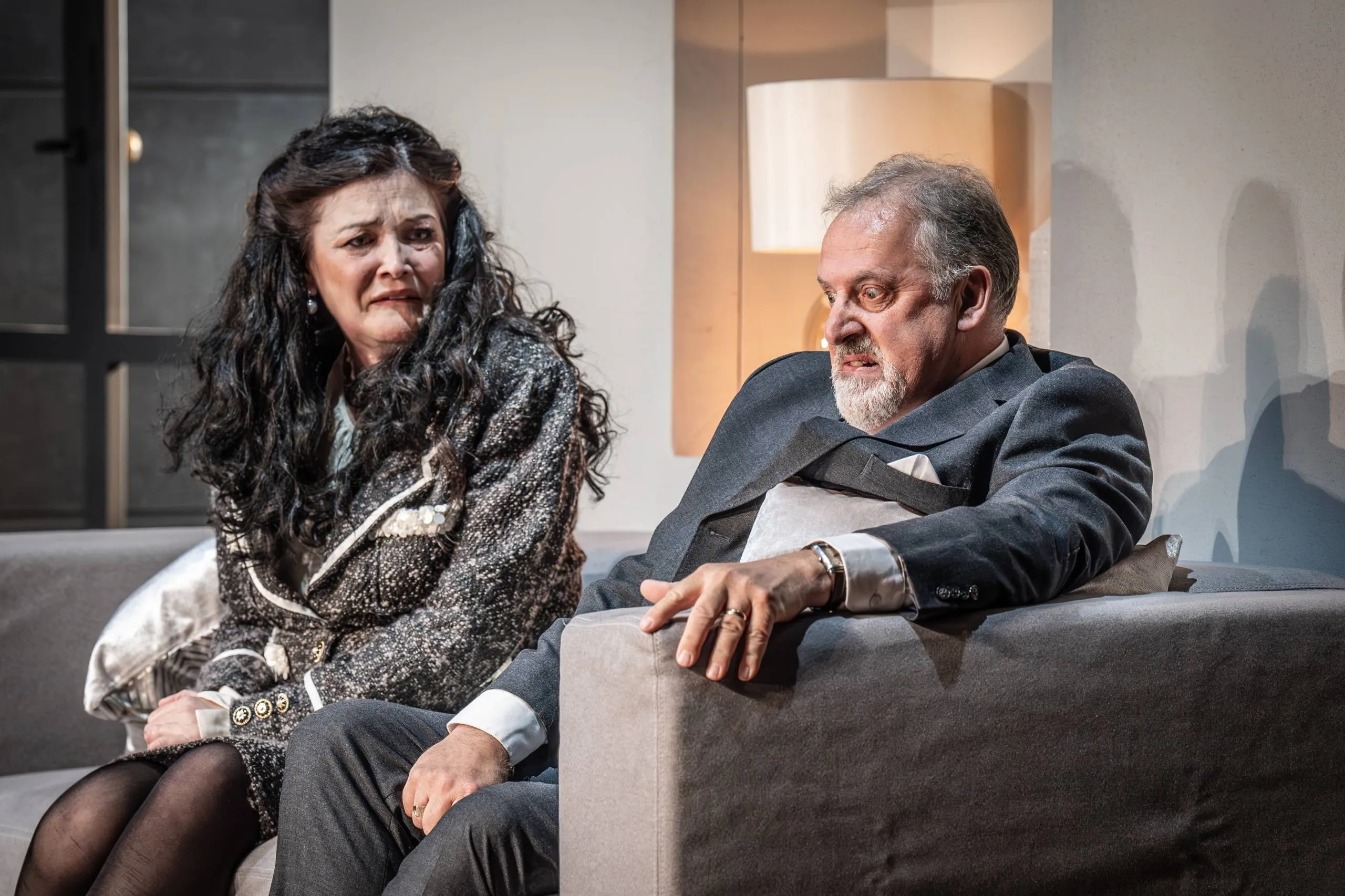 Matthew Brook seated on a sofa beside a soprano in a staged opera