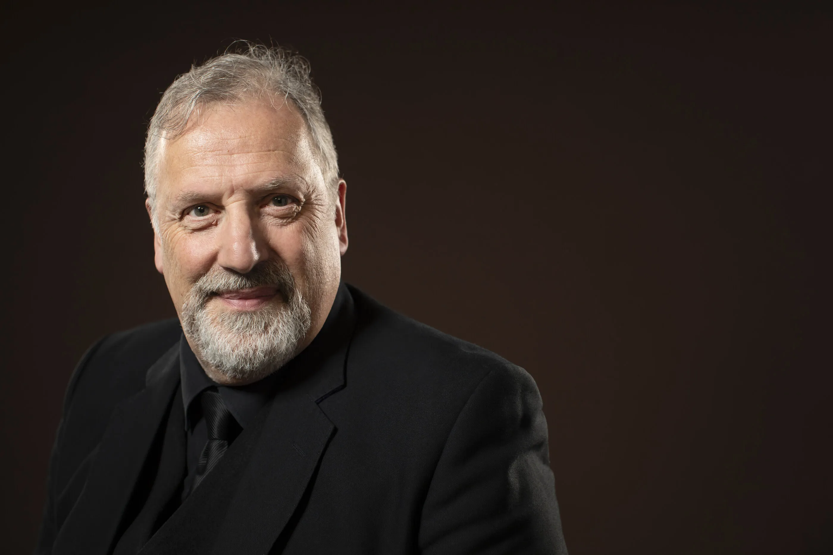 Portrait of Matthew Brook, grey-bearded, in black against a warm umber backdrop