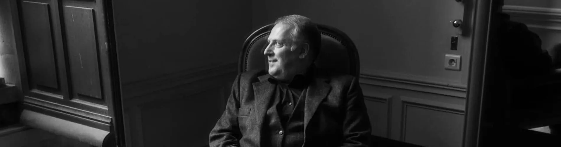 Black-and-white photograph of Matthew Brook seated in an armchair beside a panelled door, looking towards the light