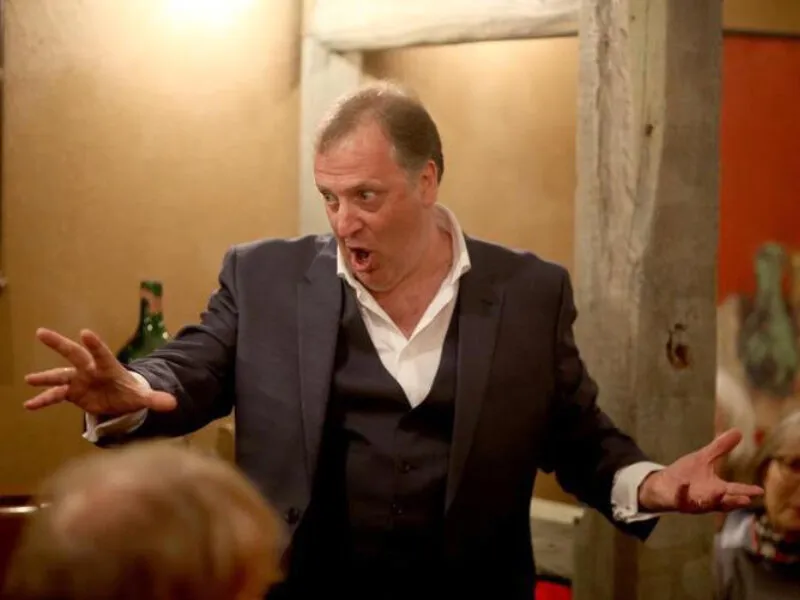 Matthew Brook in a dark suit, gesturing mid-phrase in a staged performance