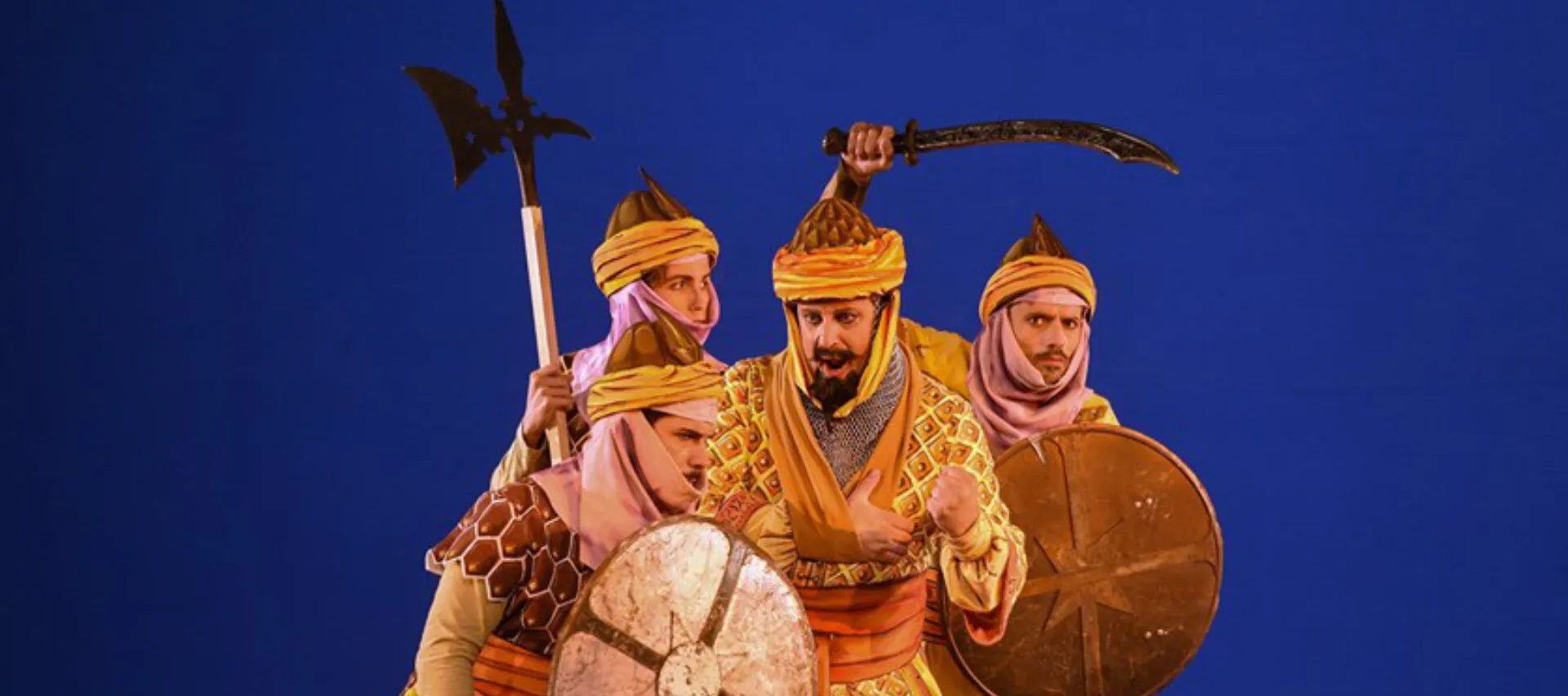 Matthew Brook in golden Ottoman-style costume with a shield, flanked by two soldiers
