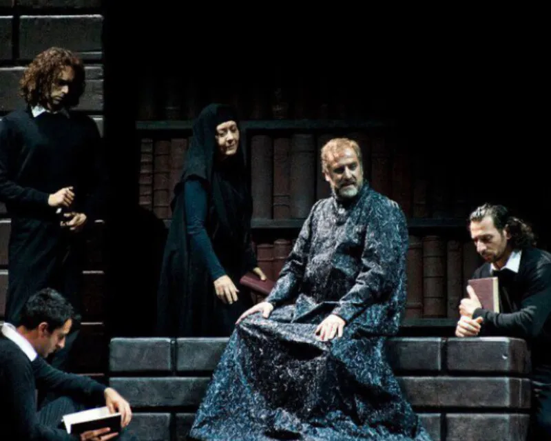 Matthew Brook in a patterned robe, seated centre stage with other singers
